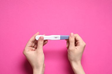 Kadının elinde pembe arka planda hamilelik testi var.