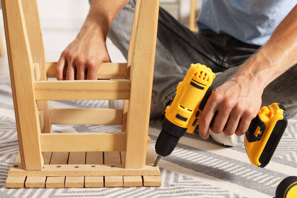 Man repairing wooden stool with electric screwdriver indoors, closeup