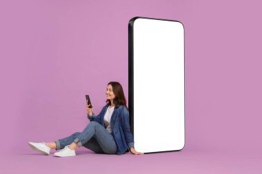 Leylak arka planında boş ekranı olan büyük bir cep telefonunun yanında otururken akıllı telefon kullanan mutlu bir kadın. Tasarım için model