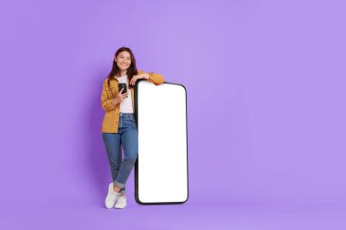 Akıllı telefonlu, mutlu, gülümseyen bir kadın. Büyük cep telefonunun yanında duruyor. Menekşe arka planında boş ekran var. Tasarım için model