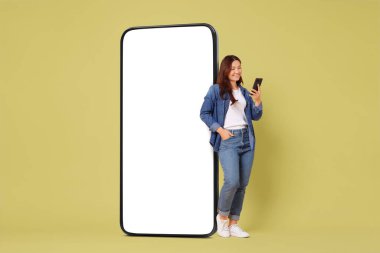 Mutlu, gülümseyen bir kadın, arka planında beyaz ekran olan büyük bir cep telefonunun yanında dururken akıllı telefon kullanıyor. Tasarım için model