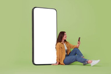 Açık yeşil arka planda boş ekranı olan büyük bir cep telefonunun yanında oturan mutlu, gülümseyen kadın. Tasarım için model