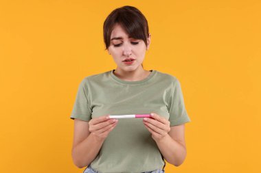 Turuncu arka planda hamilelik testi olan endişeli genç bir kadın.