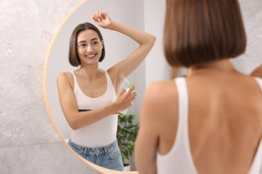Gülümseyen kadın aynanın yanında sprey deodorant sürüyor.