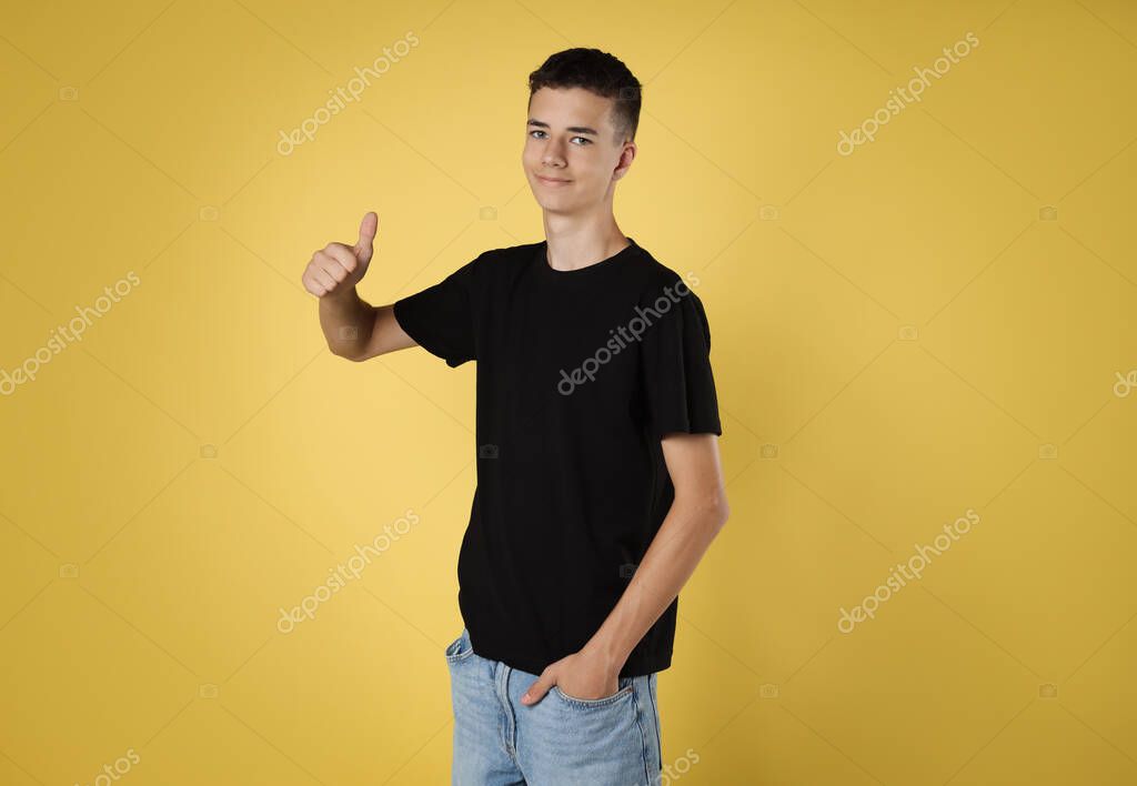 Teenage boy wearing black t-shirt and showing thumbs up on yellow background