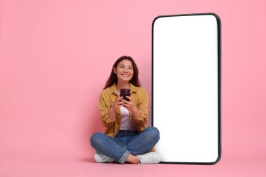 Akıllı telefonu olan mutlu bir kadın pembe arka planda boş ekranı olan büyük bir cep telefonunun yanında oturuyor. Tasarım için model