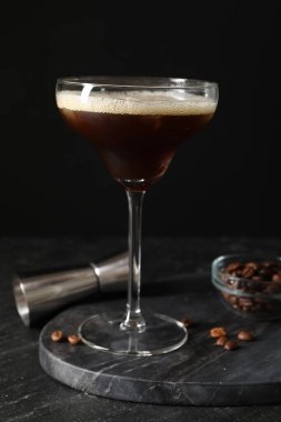 Bir bardak nefis espresso martini, jigger ve kahve çekirdekleri koyu dokulu masada, yakın plan.