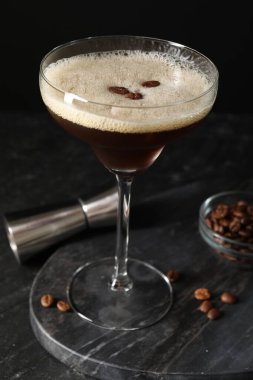Bir bardak nefis espresso martini, jigger ve kahve çekirdekleri koyu dokulu masada, yakın plan.