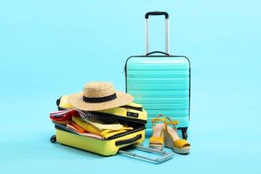 Giysileri, ayakkabıları ve açık mavi arka planda hasır şapkası olan valizler.