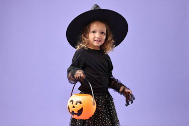 Mor arka planda cadı gibi giyinmiş balkabağı kovalı tatlı kız. Cadılar Bayramı kostümü