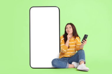Akıllı telefonu olan düşünceli bir kadın açık yeşil arka planda boş ekranlı büyük bir cep telefonunun yanında oturuyor. Tasarım için model