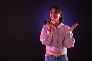 Koyu arkaplanda, neon ışıklı akıllı telefondan sesli mesaj kaydeden güzel bir kadın.