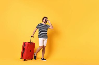 Güneş gözlüklü, bavulu turuncu arka planda yürüyen mutlu bir adam.