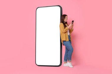 Mutlu, gülümseyen bir kadın pembe arka planda boş ekranı olan büyük bir cep telefonunun yanında dururken akıllı telefon kullanıyor. Tasarım için model