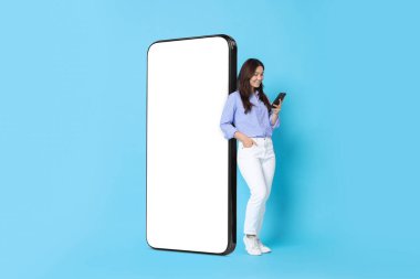 Açık mavi arka planda boş ekranı olan büyük bir cep telefonunun yanında dururken akıllı telefon kullanan mutlu bir kadın. Tasarım için model