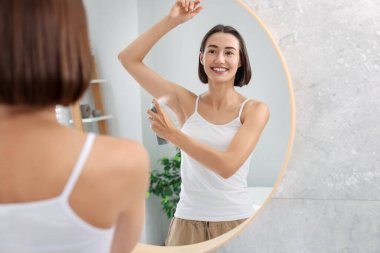 Gülümseyen kadın evde aynanın yanında sprey deodorant sürüyor.