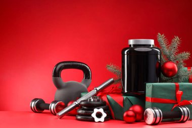 Dambıllar, kettlebell, takviyeli konteynır ve kırmızı arka planda Noel dekoru. Metin için boşluk