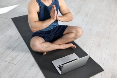 Evdeki yoga minderinde dizüstü bilgisayarın yanında meditasyon yapan bir adam.