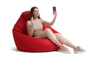 Akıllı telefonlu gülümseyen kadın beyaz arka planda kırmızı fasülye koltuğu üzerinde online toplantı yapıyor.