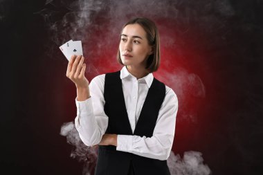 Krupiye elinde iskambil kağıtlarını koyu kırmızı arka planda dumanla tutuyor.