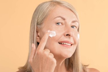 Bej arka planda yüz kremi süren son sınıf öğrencisi bir kadın.