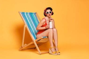 Portakal rengi arka planda katlanan sandalyede milkshake ile mayo giymiş bir kadın.