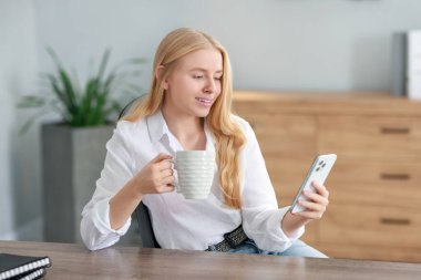 Ofiste akıllı telefon kullanan ve kahve içen gülümseyen kadın.