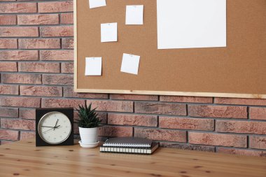 Boş kağıt notlar, çalar saat, defterler ve tuğla duvarın yanındaki ahşap masa üzerindeki ev bitkisi.