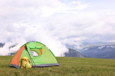 Çadır, sırt çantası, yürüyüş direkleri ve dağlardaki yeşil çimlerin üzerindeki termos.