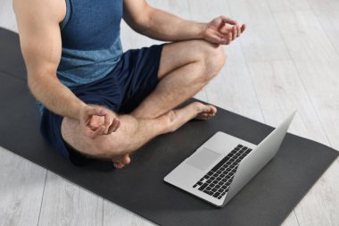 Evdeki yoga minderinde dizüstü bilgisayarın yanında meditasyon yapan bir adam.