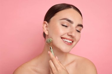Güzel genç bir kadın pembe arka planda yuvarlağıyla yüz masajı yapıyor.