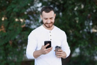 Kağıt bardaklı mutlu adam bulanık arka planda akıllı telefon kullanıyor.