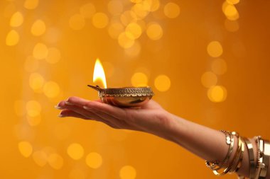 Diwali kutlaması. Elinde yanan diya lambası olan turuncu arka planda bulanık ışıkları olan bir kadın, yakın plan.