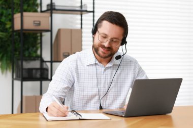 Interpreter in headset taking notes while having video chat via laptop at wooden table indoors clipart