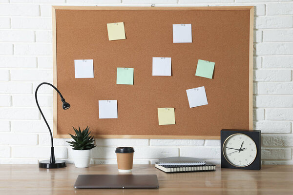 Cork board with blank paper notes, laptop, lamp, alarm clock and notebooks on wooden table near white brick wall