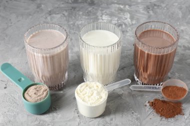 Lezzetli protein içecekleri bardaklarda ve toz dolu kepçelerle gri desenli masada, yakın plan.