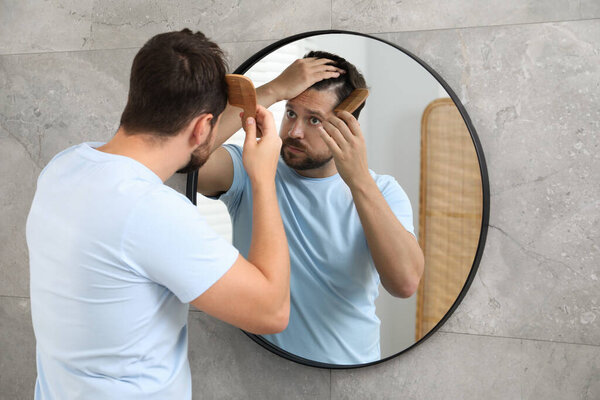 Мужчина расчесывает волосы возле зеркала в помещении. Задача об алопеции