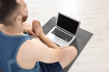 Evdeki yoga minderinde dizüstü bilgisayarın yanında meditasyon yapan adam.