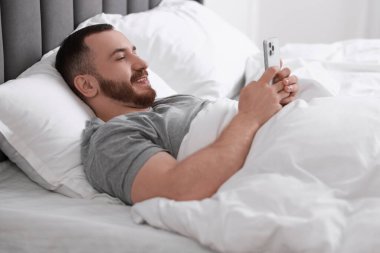 Yatakta cep telefonu kullanan gülümseyen adam.