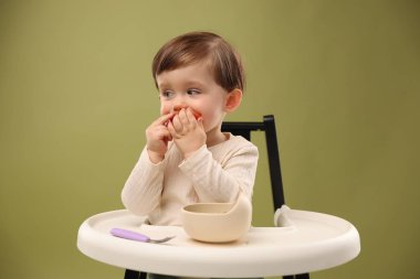 Küçük şirin bebek, zeytin arka planında yüksek sandalyedeki kaseden sağlıklı yemek yiyor.