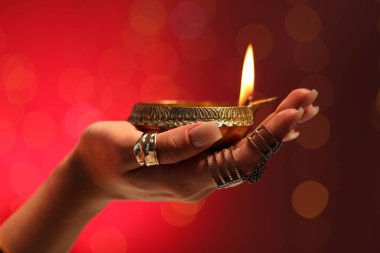 Diwali kutlaması. Kırmızı arka planda yanan diya lambasını tutan bulanık ışıklı kadın, yakın plan.