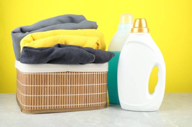 Çamaşır deterjanı şişelerde, kıyafetler gri mermer masada, sarı arka planda.