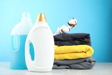 Şişelerde çamaşır deterjanı, pamuk çiçekler ve açık mavi arka planda gri mermer masa üzerindeki kıyafetler.