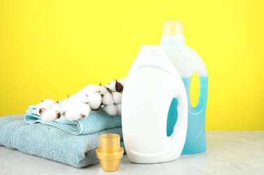 Şişe, kep, pamuk, çiçek ve havlular içinde çamaşır deterjanı. Sarı arka planda gri mermer masa.