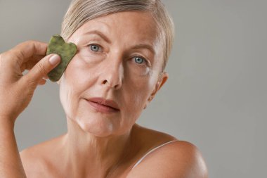 Gri arka planda gua sha aracıyla yüz masajı yapan güzel bir kadın.