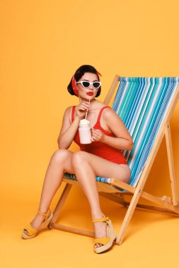 Portakal rengi arka planda katlanan sandalyede milkshake ile mayo giymiş bir kadın.