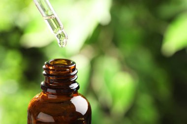 Essential oil dripping from pipette into bottle against blurred green background, closeup. Space for text clipart