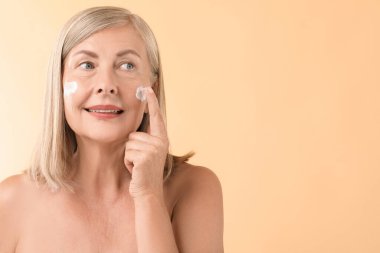 Bej arka planda yüz kremi süren son sınıf öğrencisi bir kadın. Metin için boşluk