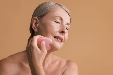 Güzel bir kadın bej arka planda gua sha aracı ile yüz masajı yapıyor, mesaj için boşluk