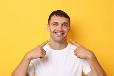 Gülümseyen adam sarı arka plandaki diş tellerine işaret ediyor.
